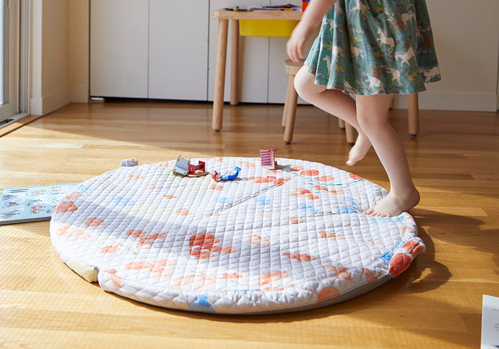 Girl's feet dancing on top of a portable cushioned baby mat with handles and the sun shining through the glass door onto the floor and her feet.