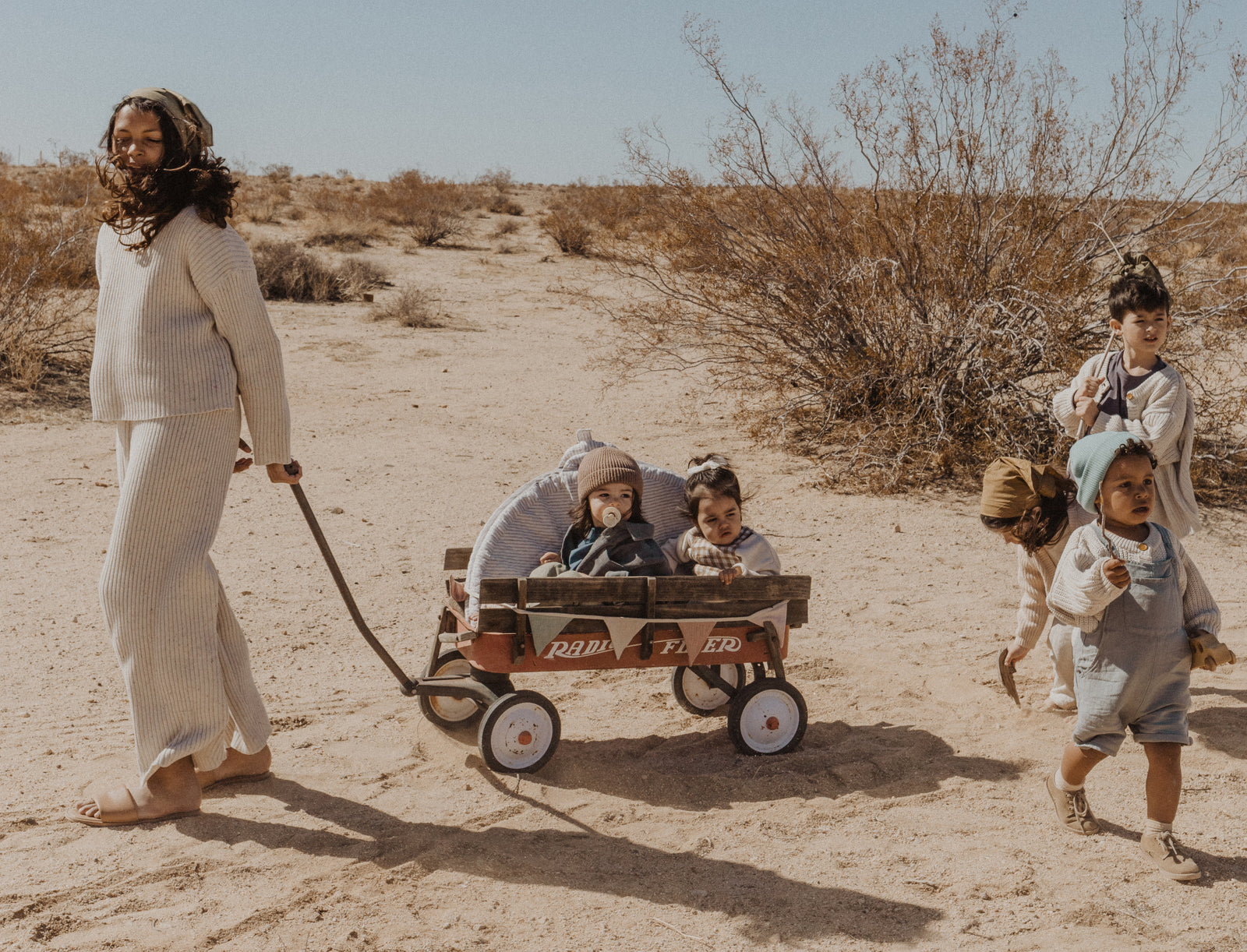 Tween girl pulling a red wagon across the desert with a portable folded baby mat and two toddlers inside and to young children playing next to them