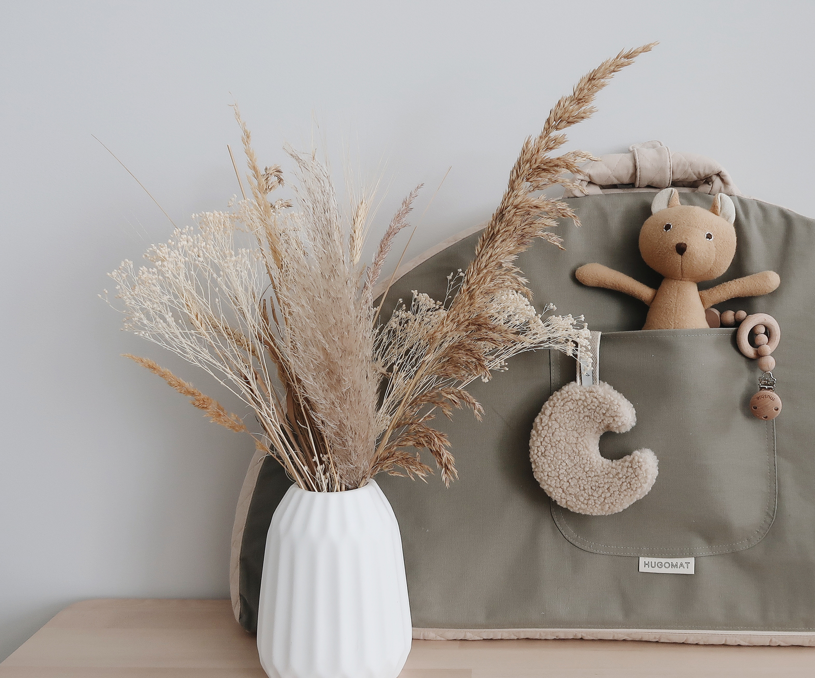Display of baby dolls and moon teether in the pocket of a folded portable baby mat with handles and a white vase with dry florals in the foreground